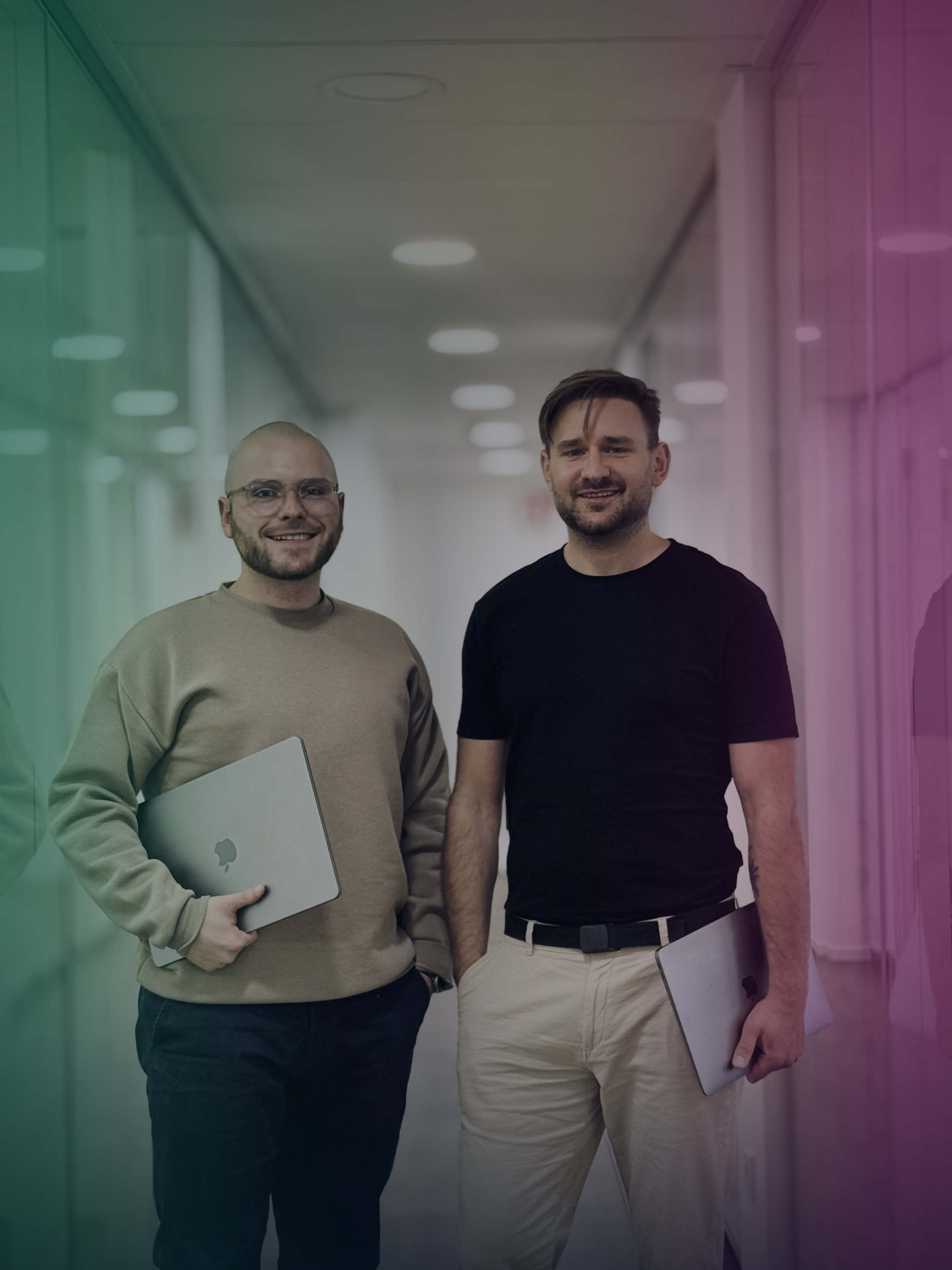 Alex and Daniel stand with laptops in an office hallway.